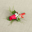 The boutonnière in this image centres on a striking magenta rose, its velvety petals arranged in a densely layered spiral that draws the gaze straight into the heart of the bloom. Around it, a collar of textured greenery frames the flower: deep green leaves provide structure, while fine, graceful blades of grass extend outward in smooth arcs, giving the design movement and an almost calligraphic elegance. Tucked between these leaves and blades are tiny sprays of white baby's breath, their miniature blossoms forming soft, clouded clusters that add both brightness and a sense of gentle nostalgia, perfect for a wedding or formal event in Mill Hill. The stems are expertly wrapped in a vivid red satin ribbon, which coils tightly and evenly from base to tip, offering a secure hold and a refined, polished finish. The satin's sheen catches ambient light, creating slim glints that echo the celebratory mood of a reception, perhaps at a venue near Mill Hill Broadway or overlooking the green spaces around Arrandene. Set against a quiet, neutral background, the colours appear rich and true: the fuchsia rose stands out vividly against the balance of green, white, and red. Imagining it pinned to a dark suit or tuxedo, you can almost sense the gentle fragrance of the rose and the crisp, fresh notes of foliage that come from flowers prepared that same day by a local Mill Hill florist. The arrangement feels intimate and expressive, the kind of small floral detail that can transform an outfit into something memorable and deeply personal.