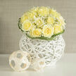 There is a deliberate clarity to this arrangement: closely gathered yellow roses form a compact dome whose ruffled centres open like soft breaths, each petal catching the light with a warm, buttery glow. Interspersed among them, clusters of tiny white gypsophila act as delicate punctuation marks-airy, cloud-like and faintly luminous against the richer rose tones. The flowers are cradled in a whitened wicker orb, a contemporary vessel whose architectural openness gives the bouquet a sense of lift and modern poise; small minimalist woven spheres nearby echo that sculptural idea and introduce a playful, artisanal note. Green blades curl with skilled restraint around the base, their looping motion adding subtle rhythm and keeping the composition visually grounded. The scent profile suggested by the image is fresh and summery: a clean rose perfume softened by the grassy, slightly mineral hint of newly cut stems-an aroma that calls to mind a walk from Mill Hill Broadway toward the common on a cool morning. As a florist in Mill Hill I value these quiet combinations that feel both intimate and refined; this design, crafted by Flowers Mill Hill, suits celebratory occasions and thoughtful congratulations alike. Light falls gently across the arrangement, revealing texture-velvety petals, the paper-thin gypsophila and the woven basket fibres-in a way that makes the whole scene feel like a small still life, tender and assured. It reads as a considered, modern classic that comforts and uplifts without ostentation.
