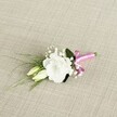 A delicately crafted wedding boutonnière lies at a soft diagonal across a pale beige, linen-like fabric, its finely woven texture echoing the quiet elegance of a Mill Hill ceremony. At the heart of the design is a single white lisianthus flower, fully open and exquisitely ruffled, its velvety petals curling in gentle waves that catch the soft, even light. Just below and to the left, two slender, creamy green lisianthus buds rest closely together, suggesting blooms that will open later in the day, much like the unfolding moments of a celebration at nearby Mill Hill Park or along The Broadway. Threaded between the central flower and buds are tiny clouds of white gypsophila, their miniature blossoms creating an airy halo that lightens and softens the piece. Behind this, a smooth, deeper green leaf provides a quiet backdrop, while several narrow, grass-like blades of foliage sweep gracefully toward the lower left corner, adding movement and a fresh, garden-picked feel. At the base, the stems are neatly gathered and bound with a vivid fuchsia pink satin ribbon, tied into a small, jaunty bow whose silky sheen stands out boldly against the neutral background. The contrast of crisp white petals, rich green leaves, and that modern flash of pink evokes a joyful, contemporary Mill Hill wedding, perhaps for a groom or groomsman arriving from a nearby church or registry office. Photographed from a slightly raised perspective, the image captures the precise craftsmanship, subtle textures, and quiet romance that make this petite floral piece ideal for a lapel on a wedding day, prom night, or any formal occasion where a single bloom can carry a world of feeling.
