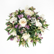 This image presents a softly rounded sympathy bouquet photographed from above, resting against a bright white background that emphasizes its calm, understated colours. At the center, plump white roses open in smooth spirals, their petals thick and silk-like to the eye, graduating from crisp white at the outer edges to warm ivory with the faintest peach undertone deep inside. Some roses are fully opened, gently cupped and welcoming, while others are still more tightly furled, adding a quiet sense of progression and time. Nestled between them are slender buds of purple alstroemeria, their surfaces shaded with an elegant gradient that begins as fresh green near the base, passes through a delicate blush of pink, and settles into muted lavender and deeper violet at the tips. A few buds show tiny cracks where the inner petals are starting to emerge, hinting at the patterned stripes they'll soon reveal. Threaded through this arrangement, tiny clusters of white baby's breath float like a fine mist, their miniature blossoms breaking up the spaces between the larger flowers and giving the bouquet a light, breathable feel. A ring of deep green foliage grounds the whole design; the leaves are broad, smooth, and slightly glossy, curving around the blooms to create a compact, harmonious shape. The overall impression is one of tenderness and restraint, the kind of floral gesture you might bring to a quiet home near Mill Hill Park or a small chapel off Flower Lane. It suggests a wish for peace and gentle remembrance, with the freshness of the stems and the soft tones of the petals offering comfort during a difficult time.