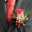A close-up, high-angle photograph shows a meticulously crafted boutonniere pinned neatly to the left lapel of a dark charcoal grey suit jacket, the kind you might see on a groom stepping out of a car near Mill Hill Broadway. At the heart of the design sits a single deep red rose, its petals tightly furled into a perfect bud that rises proudly from the fabric, catching the soft indoor light. Around the base of this rose, a halo of tiny white filler flowers, like baby's breath, creates a gentle cloud of contrast, their minute blossoms adding a delicate, almost lace-like texture. Tucked among them are several glossy, oval-shaped red berries, their sheen mirroring the richness of the rose and introducing a subtle, festive sparkle. Lush greenery frames the entire cluster, with thin, grass-like blades fanning out in slender arcs and broader, darker green leaves cupping the base, giving the arrangement structure and depth. A slim red satin ribbon is tied into a precise little bow around the stems, its polished surface echoing the rose's intensity and drawing the eye to the craftsmanship. Beneath the lapel, a vivid red tie, likely silk or satin, lies in a perfectly formed knot, reinforcing the red colour theme and giving the outfit a unified, elegant feel. A subtly patterned light shirt, perhaps a fine grey check or houndstooth, peeks out from behind the tie, adding quiet sophistication. The overall impression is of careful preparation and classic style, a polished finishing touch ideal for a Mill Hill wedding, prom, or special evening reception at a local venue, where small details convey thoughtfulness and refined taste.