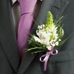 A high-angle, close-up photograph captures the upper torso of a man dressed for a formal occasion in Mill Hill, his dark charcoal grey suit jacket forming a smooth, tailored frame for an elegant wedding buttonhole. The finely cut lapel sits cleanly against a light dress shirt with a subtle grid pattern, while a sleek lavender tie drops in a precise line through the centre of the composition. Pinned to the left lapel is a carefully crafted boutonniere: a slim, pale green spire of buds, perhaps hyacinth or Muscari, rises at the top, suggesting freshness and new beginnings. Around it, open white alstroemeria blooms show off their painterly maroon streaks and soft golden stamens, their petals slightly curved and almost luminous against the darker fabric. Tiny clouds of white gypsophila, or baby's breath, weave between the larger flowers, giving the piece an airy, romantic feel, as if it were prepared moments ago in a quiet Mill Hill florist's studio before a ceremony at a local church or venue. Slender blades of grass-like foliage and broader, darker green leaves cradle the blooms, their glossy texture and rich colour adding depth and contrast. At the base, a neat lavender satin ribbon is tied into a small, symmetrical bow, echoing the tie's colour and drawing the eye to the florist's detailed handiwork. The lighting is soft and natural, highlighting the crispness of the suit and the delicate structure of each petal, creating an overall impression of calm anticipation, refined style, and the gentle excitement of a wedding morning somewhere between Mill Hill Broadway and the leafy edges of Mill Hill Park.