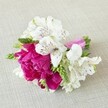 A compact, hand-crafted floral arrangement rests horizontally on a softly textured, light beige linen surface, the neutral background making every petal and ribbon glow. At the heart of the design sits a single, lavish fuchsia peony-style bloom, its many ruffled petals cupped tightly together to form a rounded, velvety focal point that feels both romantic and modern. Clustered closely around this central magenta flower are crisp white alstroemeria, or Peruvian lilies, each trumpet-shaped bloom showing six fine petals brushed with slender maroon streaks and freckles near the throat, like delicate ink strokes. Between the flowers, slim stems of fresh, vibrant green buds and spiky florets add height and texture, offering a lively contrast to the softness of the larger blossoms. The stems are neatly aligned and bound with a glossy bright pink satin ribbon, wound smoothly around the base and tied off with precision so that the colour perfectly echoes the deep fuchsia of the main flower. Gentle, even light falls across the arrangement, catching on the satin and tracing subtle shadows in the peony's folds, giving the whole piece a sense of depth and movement. The design feels like a small posy or boutonnière created for a Mill Hill wedding or prom, something you might see carried down a quiet garden path off Mill Hill Broadway or photographed in the soft light near Mill Hill Park. It conveys joyful celebration in a compact form, ideal for intimate events, thoughtful corsages, or as inspiration for DIY floral work at home in north London.