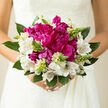 In this image, a hand-tied bridal bouquet is captured in a moment of stillness, as though the bride has paused on the way from Mill Hill Broadway to her ceremony, holding her flowers close in quiet anticipation. At the core sit opulent magenta peonies, generously full and richly textured, their many petals unfolding in soft, intricate layers that create a sense of movement even in the still photograph. The colour is a deep, romantic magenta, intense yet refined, like a burst of passion nestled at the centre of the design. Around them, white alstroemeria blooms form a graceful ring, their petals elegantly arched and delicately marked with fine burgundy striations. These markings give the impression of hand-drawn lines, adding a subtle detail that rewards a closer look and balances the peonies' boldness with quiet sophistication. Rising between the blooms are slim spikes of green buds, fresh and upright, echoing the new growth you might notice in gardens off the Ridgeway in early summer. Their presence brings both texture and symbolism, suggesting renewal and the unfolding of shared days ahead. The bouquet is enveloped in lush greenery, each leaf carefully placed to support and frame the flowers while still allowing them to look naturally gathered, as though they could have been picked from a particularly beautiful Mill Hill garden. Soft natural light washes over the arrangement, highlighting the gentle sheen on the peony petals and the translucent edges of the alstroemeria, while deeper shadows in the foliage give the bouquet depth and quiet drama. A faint sense of fragrance seems to drift from the image-fresh, green, and delicately floral-conjuring the calm of a cool wedding morning in Mill Hill. The result is an arrangement that feels both intimate and celebratory, ideal for marking a milestone with grace and feeling.