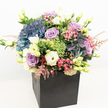 Nestled within a modern matte black square box, this stunning floral arrangement bursts with the lively colour and abundant texture of a Mill Hill spring garden. Voluminous rounds of periwinkle and blue hydrangea take centre stage, their plush petals blending seamlessly with the layered lavender and blush pink roses that swirl gently throughout. Creamy white and sunny yellow lisianthus infuse the composition with a soft radiance, echoing the bright freshness found in Mill Hill's local parks. Playful tufts of lime-green dianthus, reminiscent of mossy lawns, punctuate the bouquet, their unique forms adding intrigue and energy. Delicate sprays of feathery pink astilbe lend lightness and airy grace, while vibrant green foliage and inviting new buds spill naturally around the blooms. The balanced harmony of cool and warm tones, vivid flora, and subtle greenery creates a refined yet approachable luxury-perfect for milestone celebrations, heartfelt thanks, or simply bringing a sense of natural joy to homes and offices in Mill Hill. Surrounded by crisp white light, each flower shines, expertly arranged to provide beauty from every angle, and reflective of the artistry and passion of a true local florist.