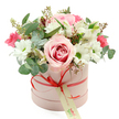 A beautifully composed hatbox bouquet is photographed against a clean white background, making its pastel tones and rich textures stand out with clarity. The box itself is a soft, powdery pink cylinder with a matte finish, giving it a modern, understated elegance. Around the lower third of the hatbox runs a slim coral-red satin ribbon, neatly tied into a small bow at the front, its smooth surface catching the light in gentle highlights. Hanging from this bow is a rectangular cream gift tag adorned with green script, suggesting a handwritten dedication ready to be read by someone in Mill Hill as they receive a surprise delivery at their door. From the open top of the box, the bouquet rises in a full, domed shape, creating a feeling of abundance without losing refinement. At the centre, a fully open bi-colour rose in pale blush and deeper pink draws immediate attention, each velvety petal curling outward in soft spirals. Around this focal bloom cluster crisp white daisy-like chrysanthemums, their bright green centres sparkling against the white petals like tiny bursts of freshness. Slender white alstroemeria blooms with characteristic speckles thread delicately through the arrangement, while clusters of smaller deep pink or fuchsia flowers, possibly spray roses or lisianthus, add pockets of vivid, romantic colour. The bouquet is generously laced with foliage: cool, rounded eucalyptus leaves in silvery green, deeper green leaves for structure, and a few finer stems topped with tiny buds that lend a touch of natural movement. The lighting is soft and even, revealing every texture from the satin ribbon to the slight ruffles on the chrysanthemum petals. The overall effect is of a thoughtful, luxurious hatbox arrangement, perfectly suited for delivery across Mill Hill for birthdays, anniversaries, Mother's Day, or any occasion when a graceful floral gesture is needed.