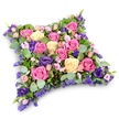 The photograph shows an elaborate sympathy cushion, a floral pillow meticulously arranged to convey comfort and respect, much as you might see at a funeral service in Mill Hill. The tribute takes the form of a low, rectangular cushion with gently softened corners, its surface completely covered in flowers so that it appears richly padded and inviting to the eye. Large roses in vivid fuchsia, soft pink, and creamy pale yellow dominate the arrangement, many fully opened so their layered petals form lush, velvety spirals that suggest both beauty and tenderness. Threaded through these are deep royal purple lisianthus blooms, their ruffled petals and slightly cupped shape adding a touch of luxury and contemplative depth. Between the statement flowers, clouds of tiny lavender-hued filler blossoms-akin to statice or limonium-provide fine detail and a misty veil of gentle colour, while miniature pink spray roses peek out, bringing a sense of closeness and affection. The whole composition is supported by an abundance of greenery: rounded eucalyptus leaves in silvery green soften the look and introduce a cool, tranquil tone, while darker, fern-like foliage adds structure and a hint of natural movement, echoing the greenery found in the parks and gardens around Mill Hill. Resting on a plain white background, the cushion casts a discreet shadow that defines its plump profile without distracting from the flowers. The overall impression is one of peace, remembrance, and quiet care-a floral pillow designed to lie beside a loved one in their final farewell, offering a visual message of sympathy, love, and hope for restful sleep.