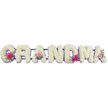 This image shows a loving funeral tribute that spells out GRANDMA in large, substantial letters, each one completely blanketed in fresh white chrysanthemums. The blooms are set so closely together that they form a smooth, padded surface, their overlapping petals creating a soft, cloud-like texture across the word. A slim trim of lilac satin ribbon runs along the outer edge of each letter, forming a continuous border that gently catches the light and adds a quiet, feminine accent to the clean white flowers. At three carefully chosen points-the first G, one of the middle As, and the final A-the uniform field of white is broken by small, rounded posies of brighter blossoms. These little clusters feature vivid pink gerberas with open, daisy-like faces, softer pink roses, and sprigs of glossy greenery, lifting the design with a hint of colour and natural movement. The tribute is lit evenly, with no strong shadows, allowing every floret, ribbon edge, and leaf vein to be clearly seen against the neutral backdrop. It has the look of a piece freshly prepared in a Mill Hill florist studio, perhaps by the team at Flowers Mill Hill, ready to be taken to a farewell service at a nearby chapel or crematorium. The overall impression is both tender and dignified: a simple, readable word made special through careful floristry, conveying the warmth, steadiness, and enduring love that the title Grandma holds for so many families in and around Mill Hill.