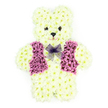 The photograph captures a three-dimensional teddy bear constructed entirely from fresh flowers, a touching memorial tribute designed with both artistry and emotion. The bear is positioned upright, facing forward, with rounded ears, a full head, curved arms, and short legs that rest in a seated pose, echoing the shape of a traditional soft toy. Every surface of its body is covered in dense, creamy white chrysanthemums, the blooms arranged so closely together that no stems or gaps are visible. This creates an unbroken layer of petals that looks as soft as plush fabric, subtly shifting in tone from pale ivory to gentle cream under the light. The focal point of colour is a tailored-looking waistcoat laid across the teddy's chest, crafted from a mix of deep purple and pastel lilac flowers. Darker, amethyst petals trace the edges and suggest lapels, while the inner area is filled with lighter lilac petals that appear slightly ruffled, adding depth and a sense of textile-like richness. At the neck, a delicate bow made from sheer lavender mesh is tied neatly, its gossamer loops and trailing ends resting lightly against the chrysanthemum fur. The bear's expression is formed from small, dark floral elements used for the eyes and nose, with a gentle, curved mouth giving an impression of calm, affectionate warmth. Though the photograph is still, you can almost imagine the quiet, fresh scent of the blooms in a chapel or family home in Mill Hill, offering subtle comfort. This floral teddy bear is a deeply personal choice for a funeral or remembrance service, conveying innocence, love, and the enduring tenderness of cherished memories.